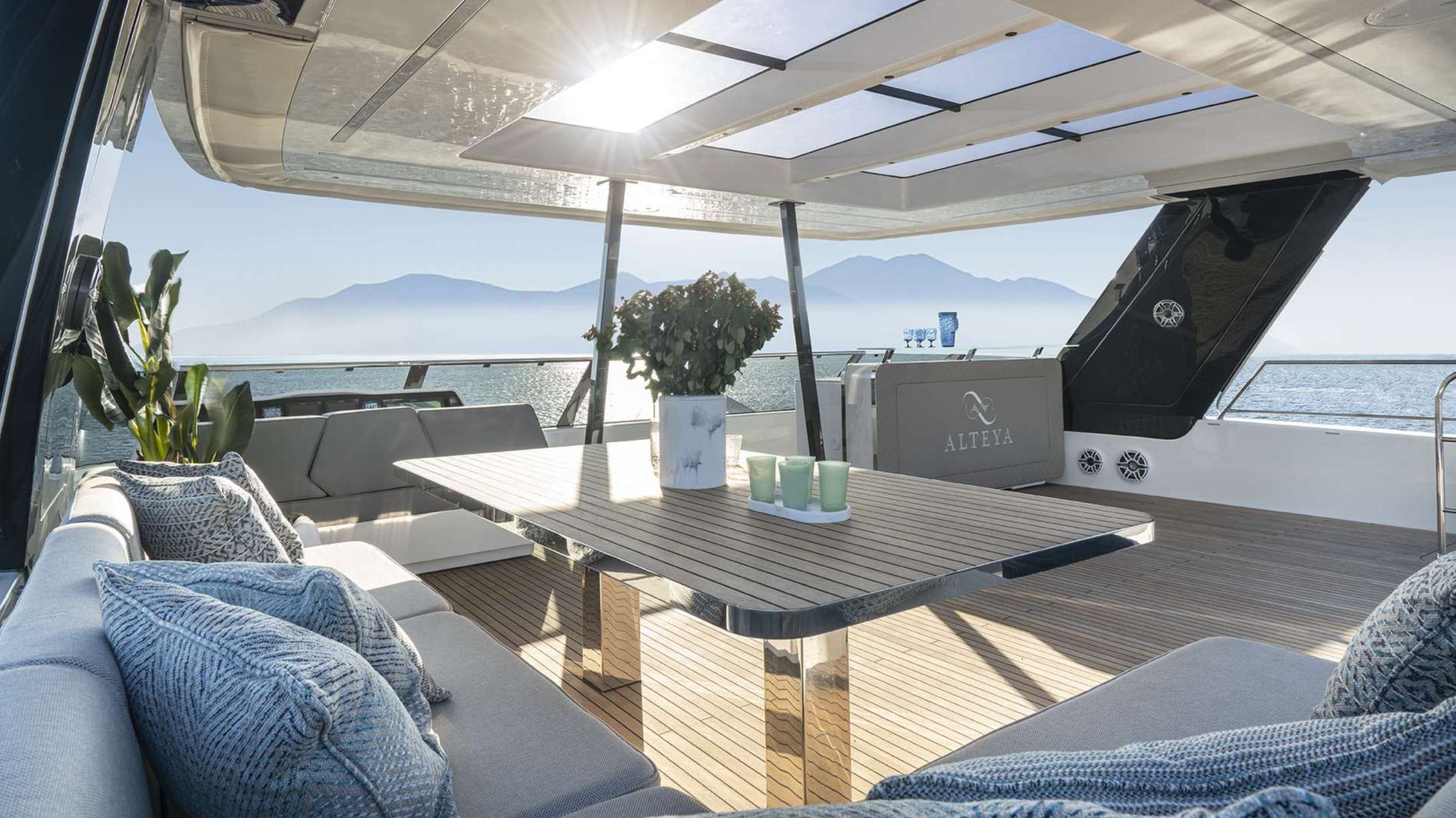 Luxurious yacht deck with cushioned seating, a wooden table topped with plants and candles, and breathtaking views—perfect for those seeking the ultimate yacht charter experience amid calm waters and distant mountains under a sunny sky.