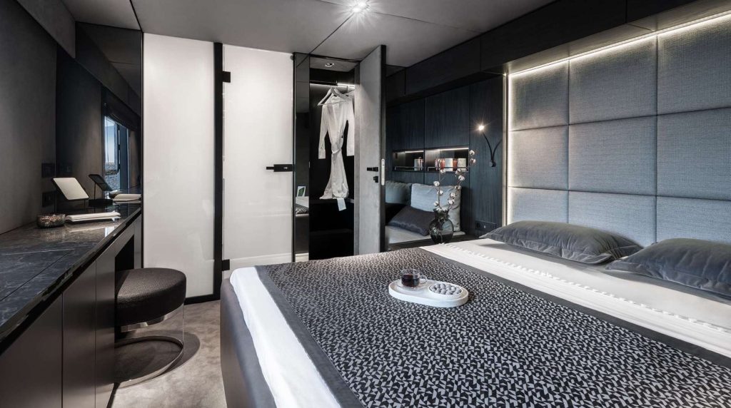 A modern, minimalist bedroom with a double bed, gray padded headboard, sleek black furniture, a vanity with a round stool, and an open wardrobe displaying a white dress—perfect comfort after a luxurious yacht charter adventure.