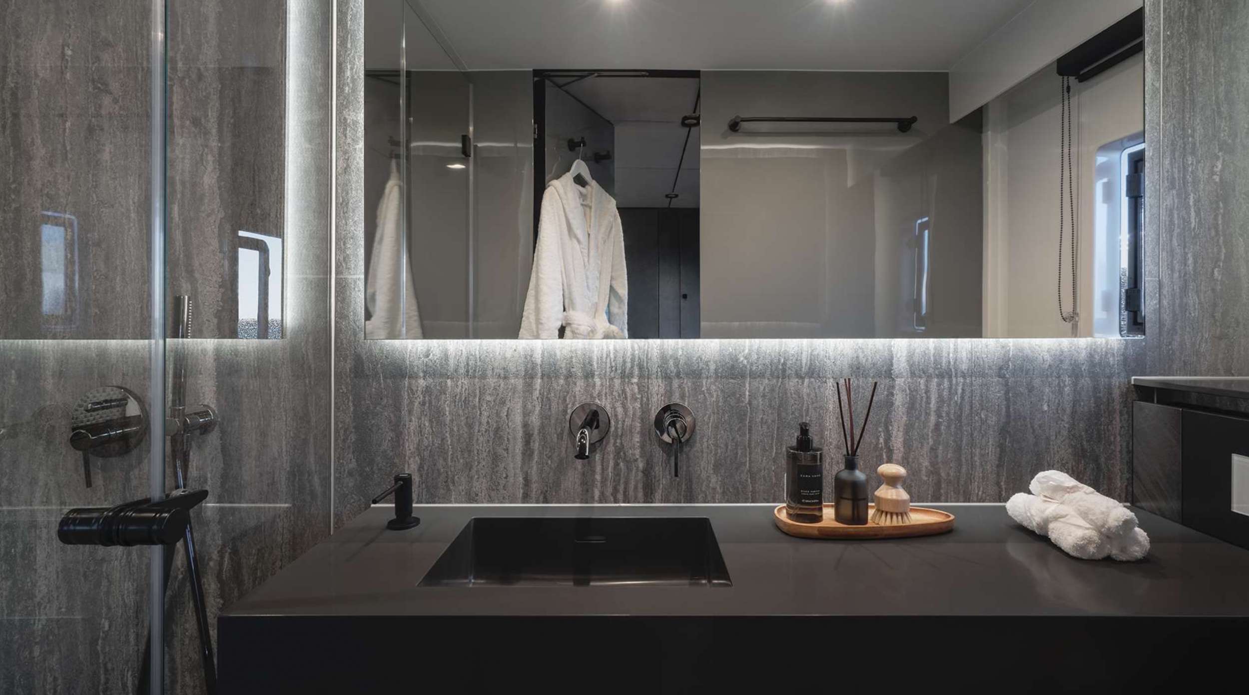 Modern bathroom with dark stone walls, black sink, large mirror, glass shower door, and a tray holding toiletries and rolled towels; a white robe hangs in the reflection—perfect luxury for a yacht charter experience.