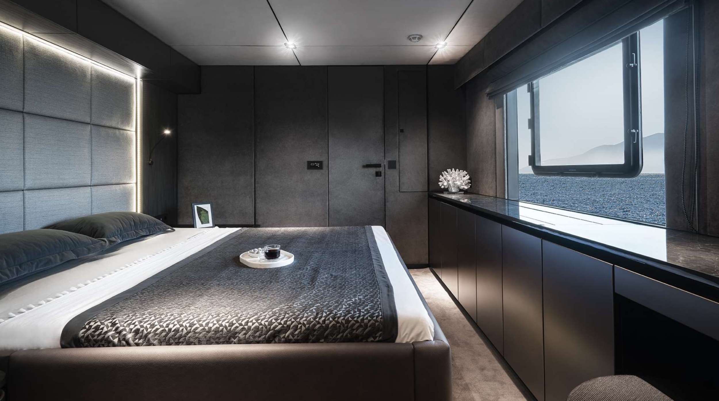 Modern bedroom with a large bed, gray walls, and minimal decor. A wide window reveals sea and mountain views dotted with yachts for charter. Built-in cabinets run along the window, and soft lighting creates a calm, luxurious atmosphere.