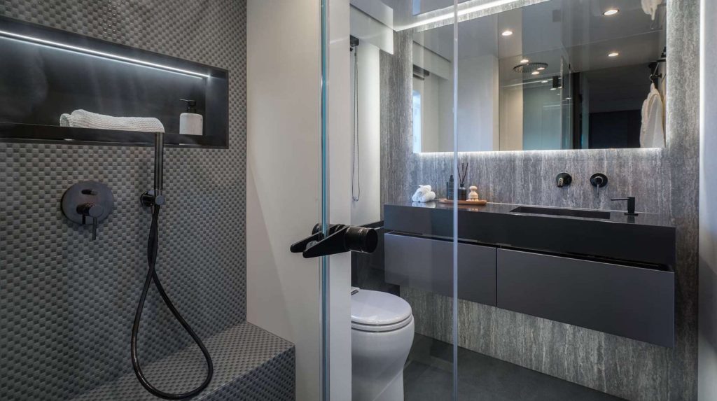 Modern bathroom with textured gray shower wall, glass door, built-in shelf holding a towel and bottle, black fixtures, floating vanity with large mirror, and a white toilet—luxury you’d expect on upscale yachts for charter.