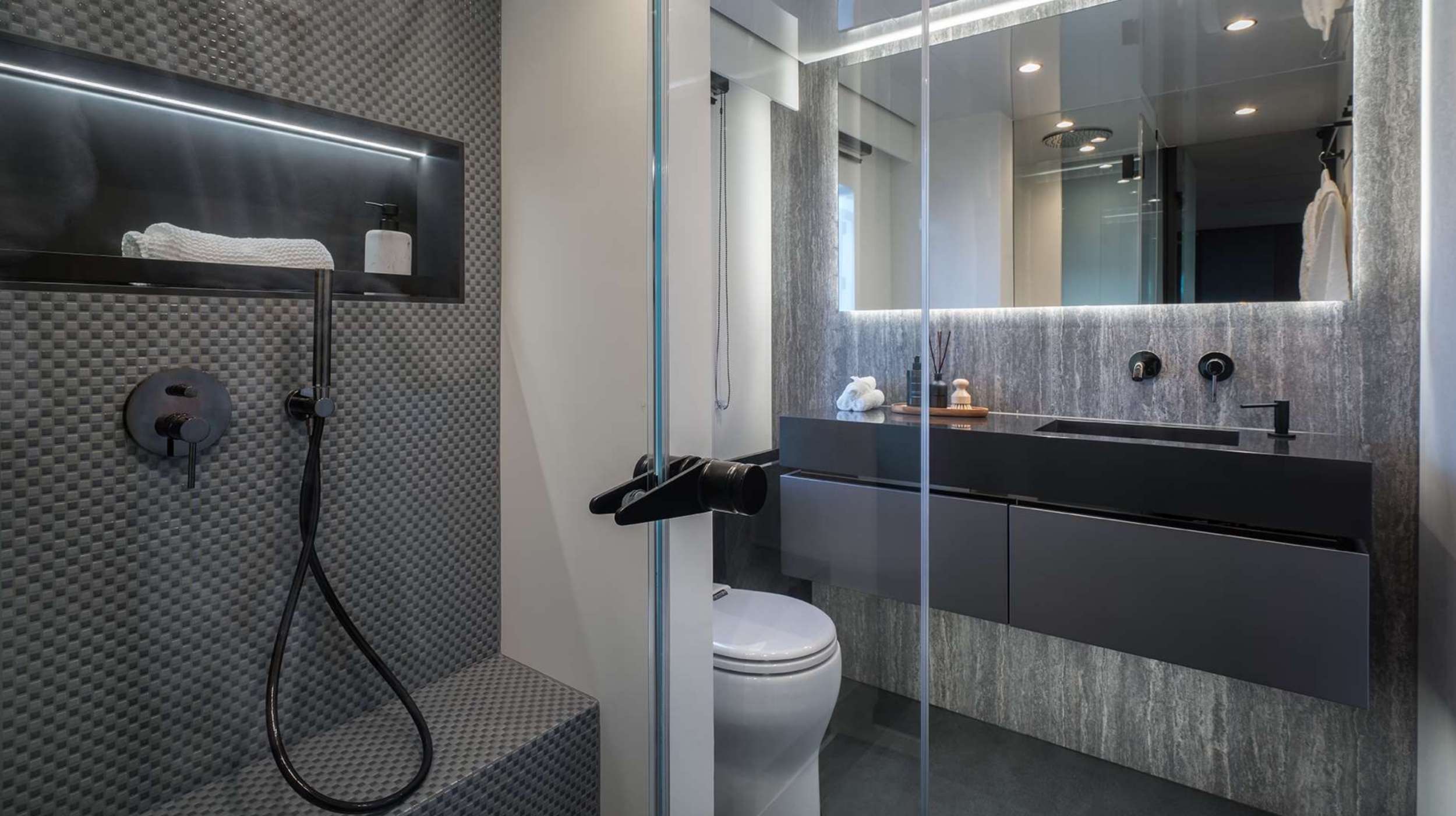 Modern bathroom with textured gray shower wall, glass door, built-in shelf holding a towel and bottle, black fixtures, floating vanity with large mirror, and a white toilet—luxury you’d expect on upscale yachts for charter.
