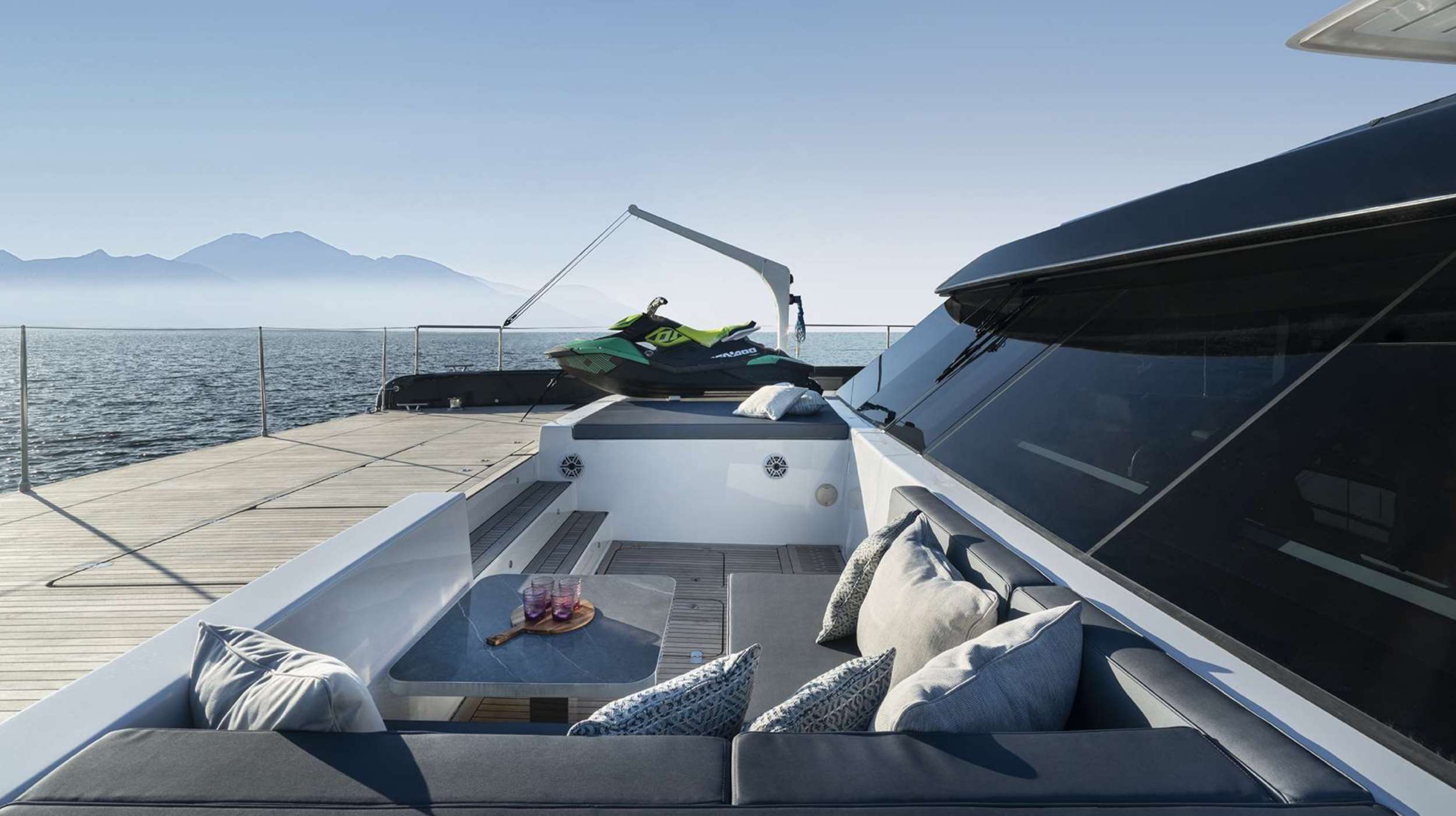 A modern yacht deck with cushioned seating, a small table holding drinks and grapes, and a green jet ski at the back—perfect for those seeking a relaxing yacht charter. Calm water and distant mountains stretch out under a clear sky.