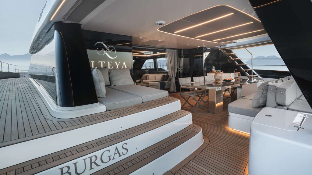 Luxury yacht deck with modern outdoor seating, dining table, and cushions under a sleek canopy with LED lighting. Ideal for a yacht charter, ITEYA displays BURGAS on the steps, with ocean and mountains in the background.