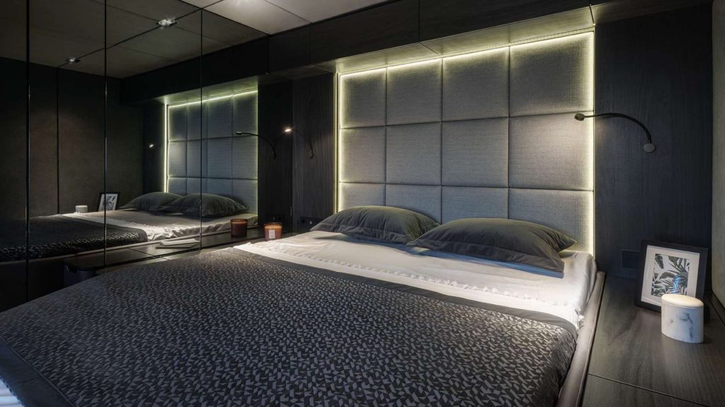 A modern bedroom with a large bed, gray padded headboard with built-in LED lighting, two black pillows, mirrored walls, and minimal decor reminiscent of luxury yachts for charter—featuring a framed photo, candle, and small lamp on a dark bedside table.