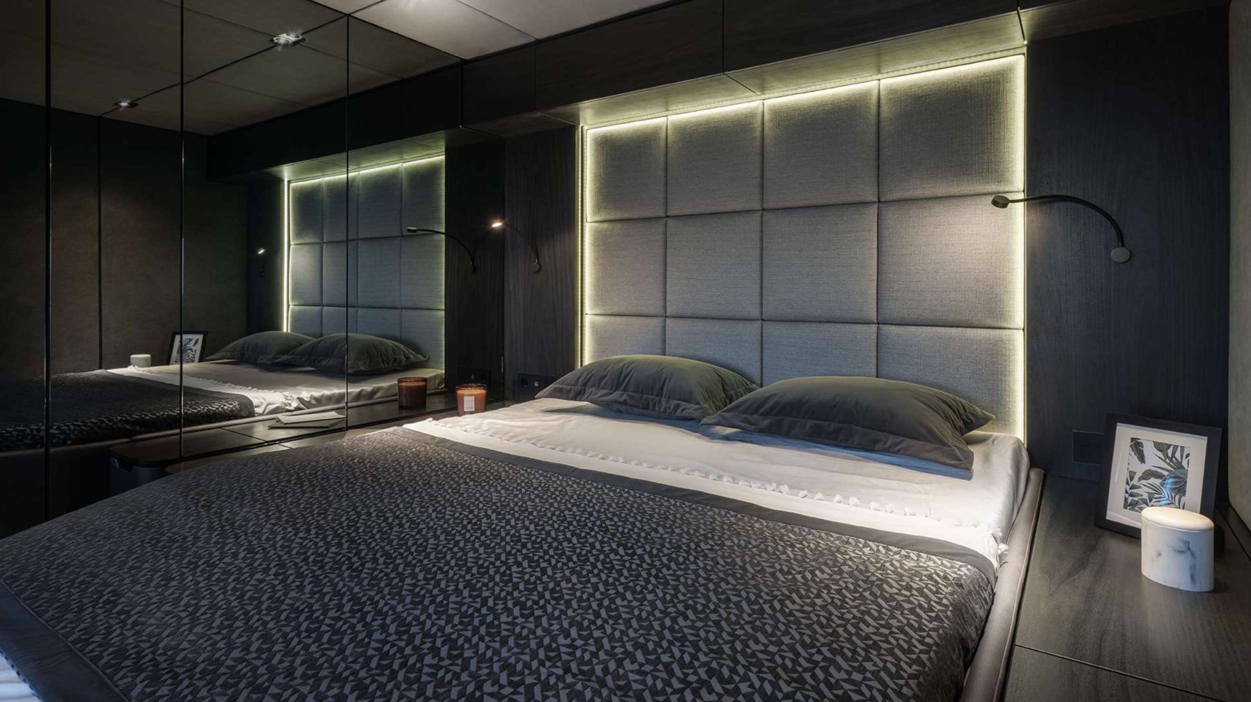 A modern bedroom with a large bed, gray padded headboard with built-in LED lighting, two black pillows, mirrored walls, and minimal decor reminiscent of luxury yachts for charter—featuring a framed photo, candle, and small lamp on a dark bedside table.