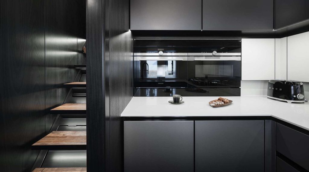 A modern kitchen with dark cabinets, wood shelves, a white countertop, and sleek finishes—perfect for a yacht charter experience where style meets functionality. Enjoy coffee and cookies as you relax in this minimalist galley.