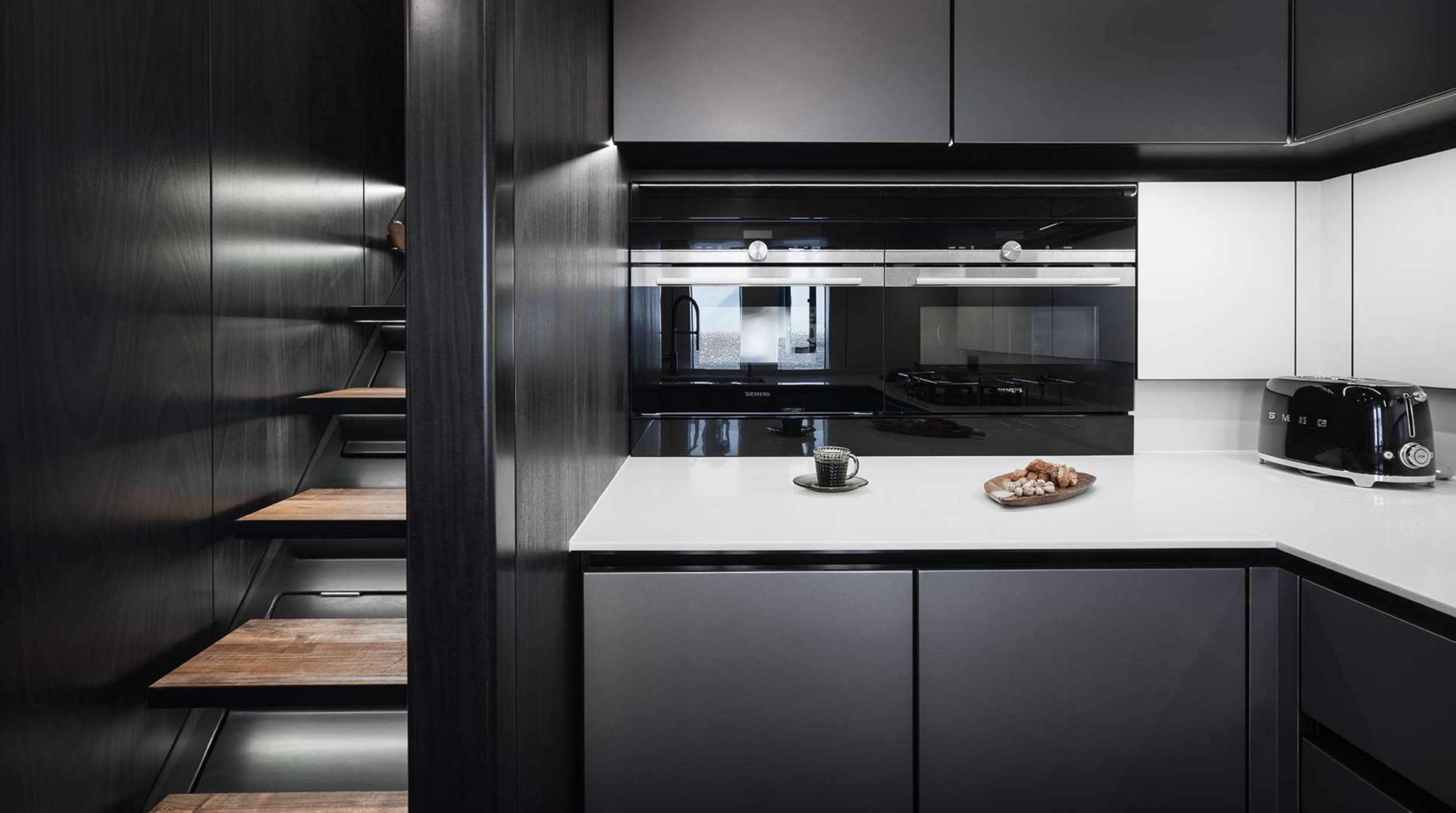 A modern kitchen with dark cabinets, wood shelves, a white countertop, and sleek finishes—perfect for a yacht charter experience where style meets functionality. Enjoy coffee and cookies as you relax in this minimalist galley.