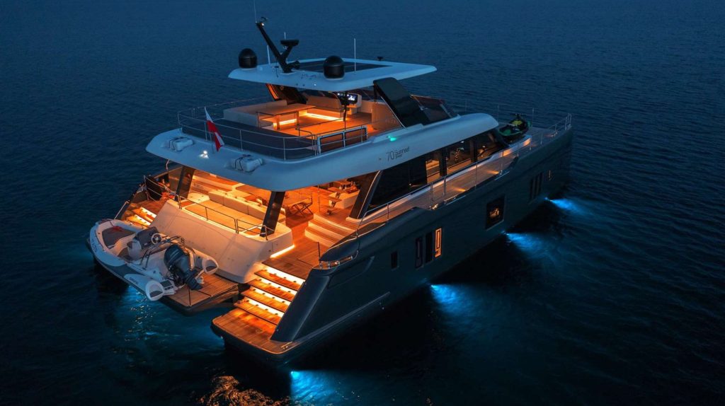 A luxury yacht available for charter glows with blue underwater lights at dusk. Warmly lit interiors and deck offer inviting outdoor seating, with a small boat and jet ski at the back, all surrounded by calm, dark water.