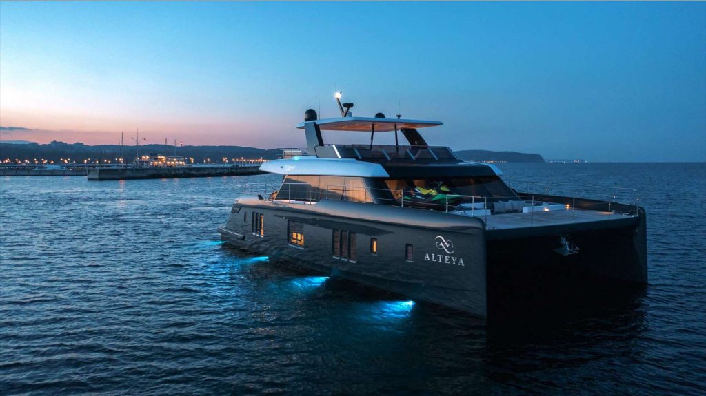 A modern yacht named Alteya, available for yacht charter, glides near the coastline at dusk, its blue underwater lights glowing as city lights and distant hills form a stunning backdrop.