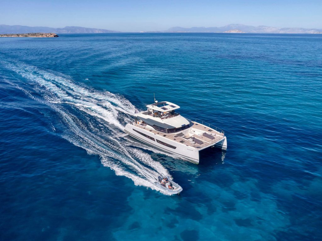 A large white catamaran yacht sailing on clear blue water, followed closely by a small motorboat. Rocky shores and distant mountains are visible under a clear sky.