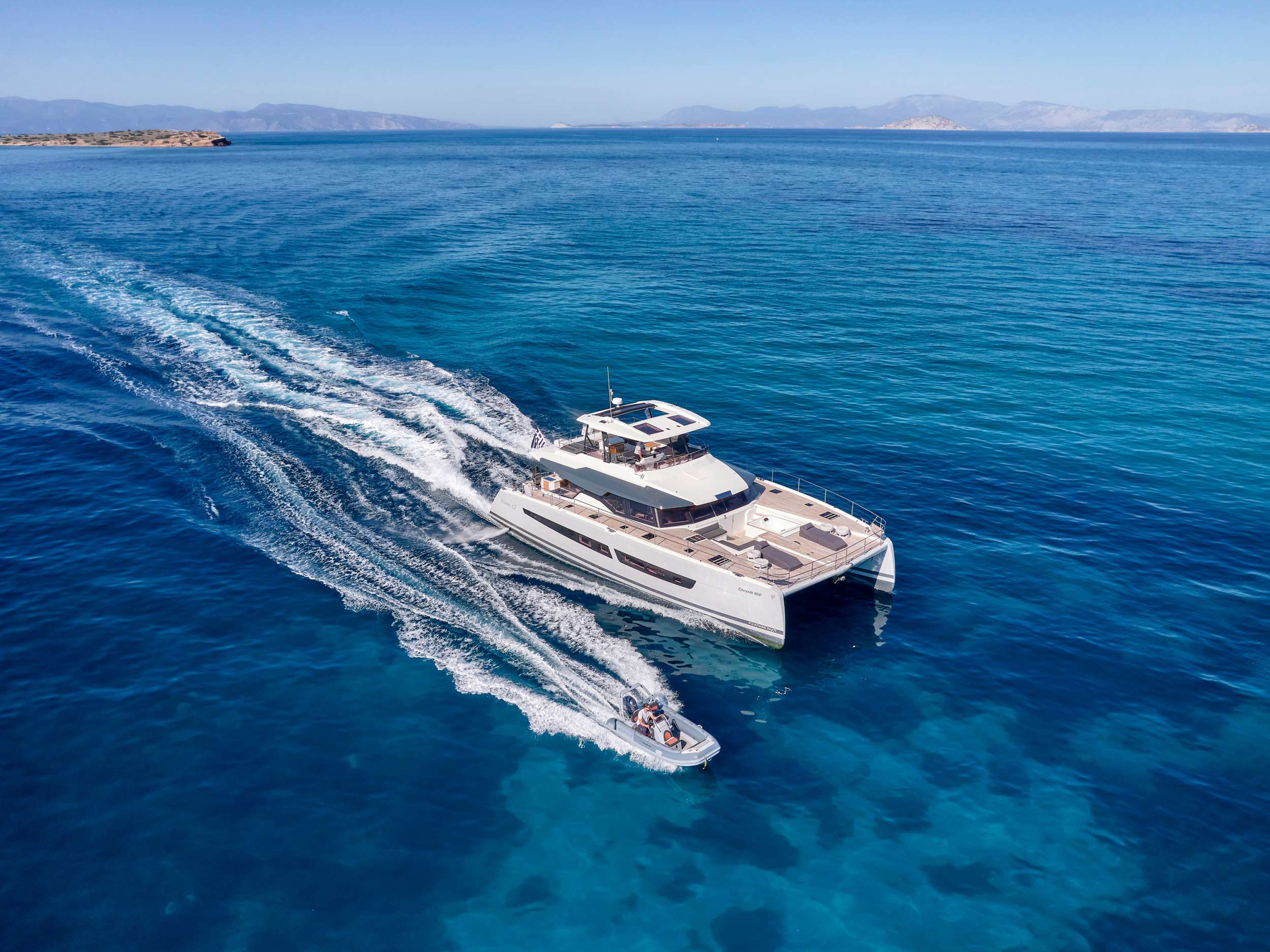 A large white catamaran yacht sailing on clear blue water, followed closely by a small motorboat. Rocky shores and distant mountains are visible under a clear sky.