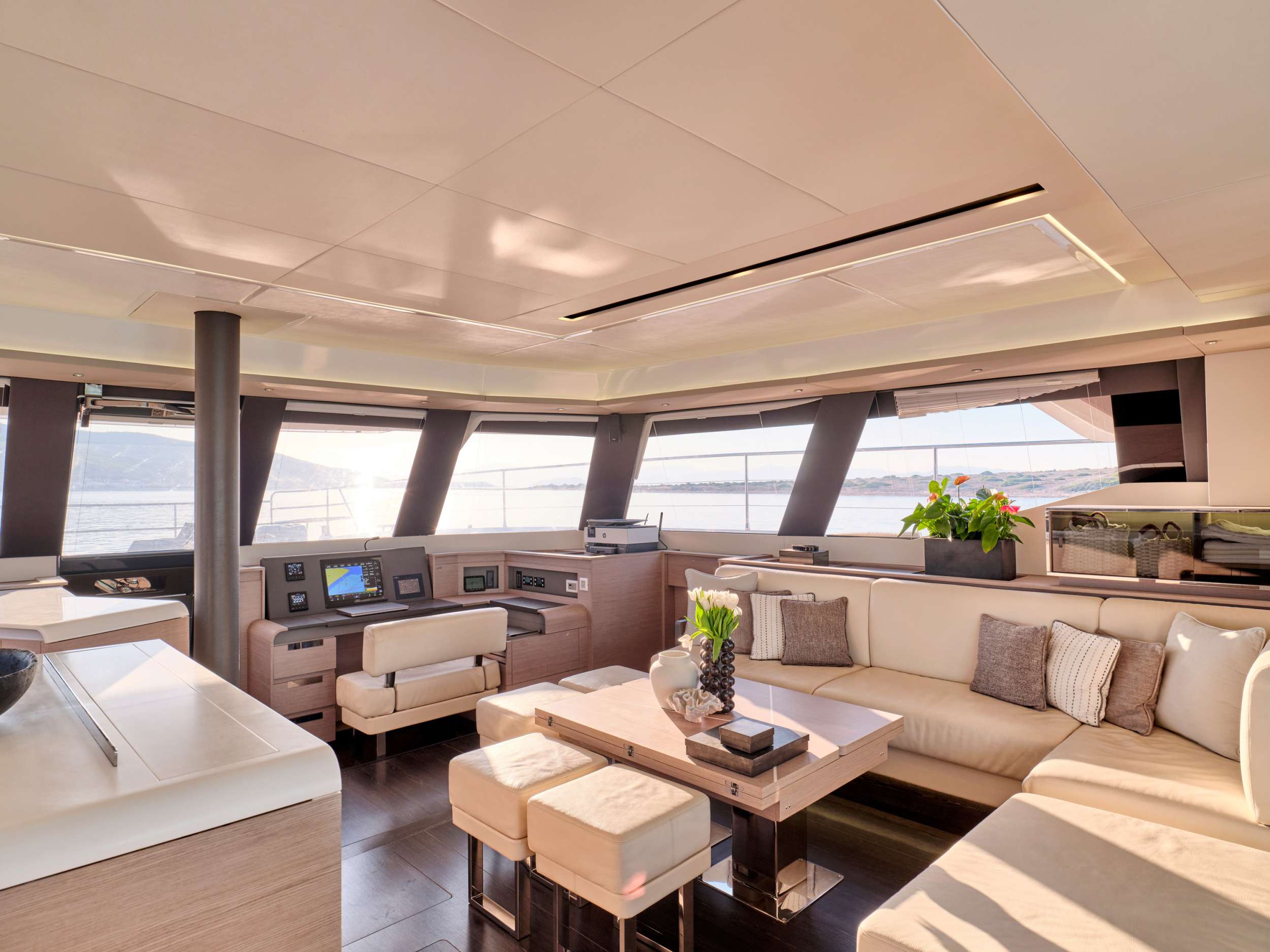 Modern yacht interior with large windows, cream-colored seating, a wooden table set with decorative items, throw pillows, and potted plants. Sunlight streams in, revealing water and distant land outside.