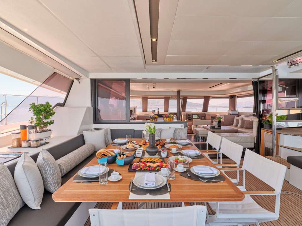 A dining area on a yacht with a wooden table set for breakfast, surrounded by white chairs and gray cushioned benches. The table is filled with pastries, fruit, and drinks. The yacht interior is modern and spacious.