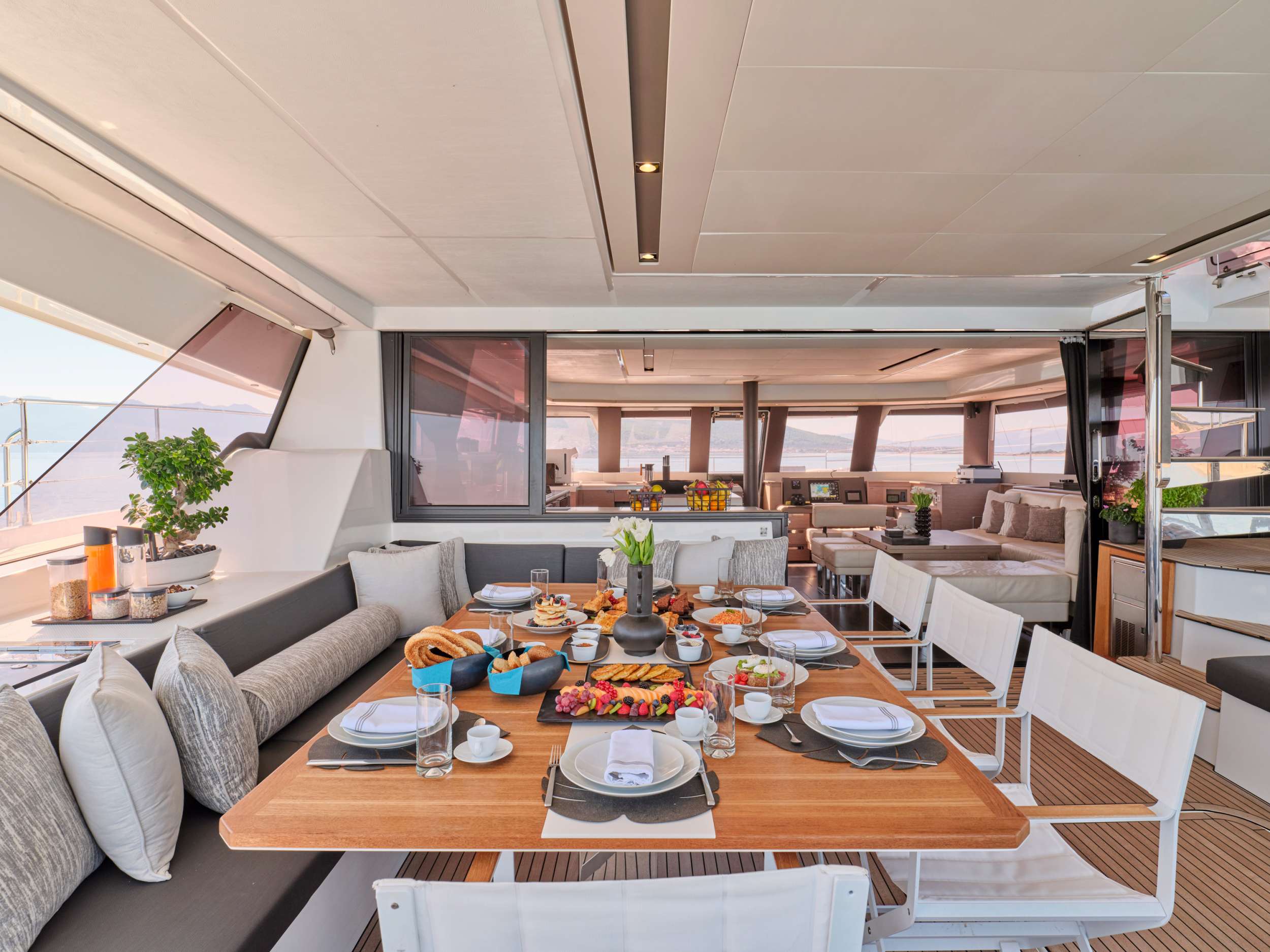 A dining area on a yacht with a wooden table set for breakfast, surrounded by white chairs and gray cushioned benches. The table is filled with pastries, fruit, and drinks. The yacht interior is modern and spacious.