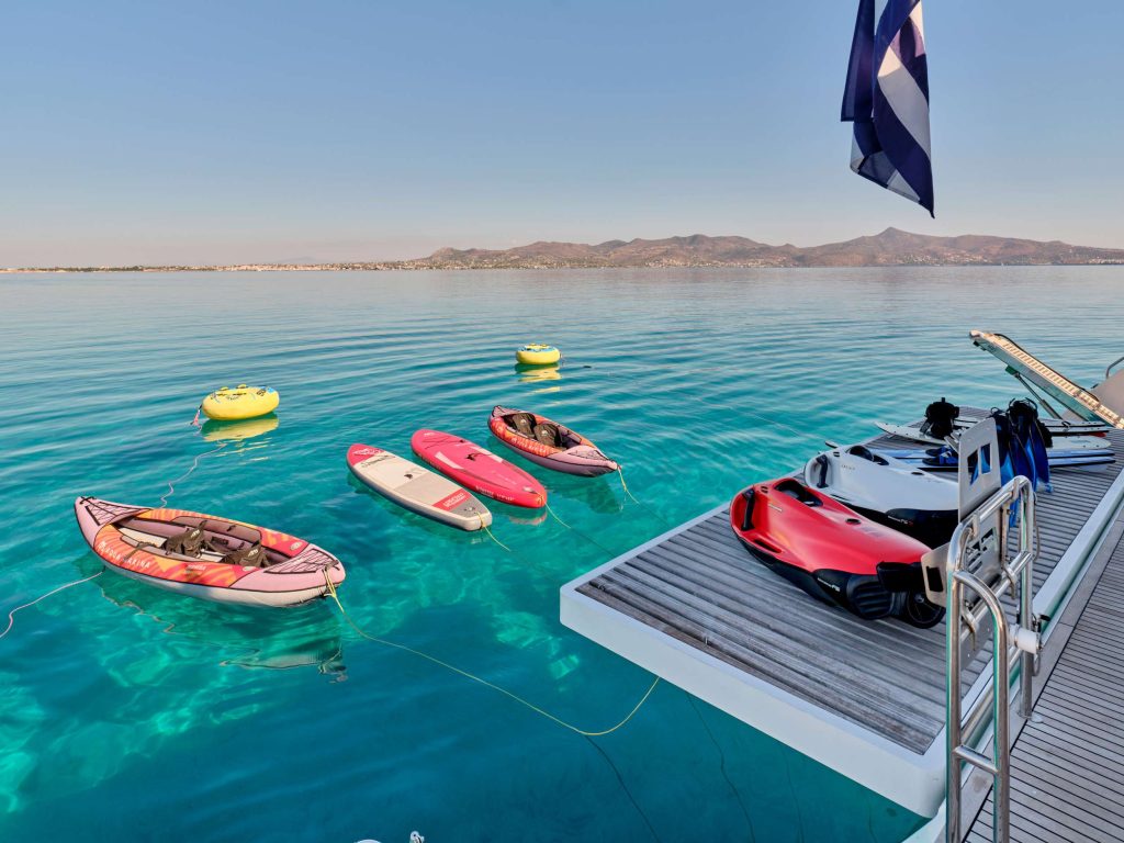 A deck overlooking crystal clear blue water with inflatable boats, paddleboards, and water toys floating nearby; a Greek flag waves on the right, with distant hills visible across the calm sea.