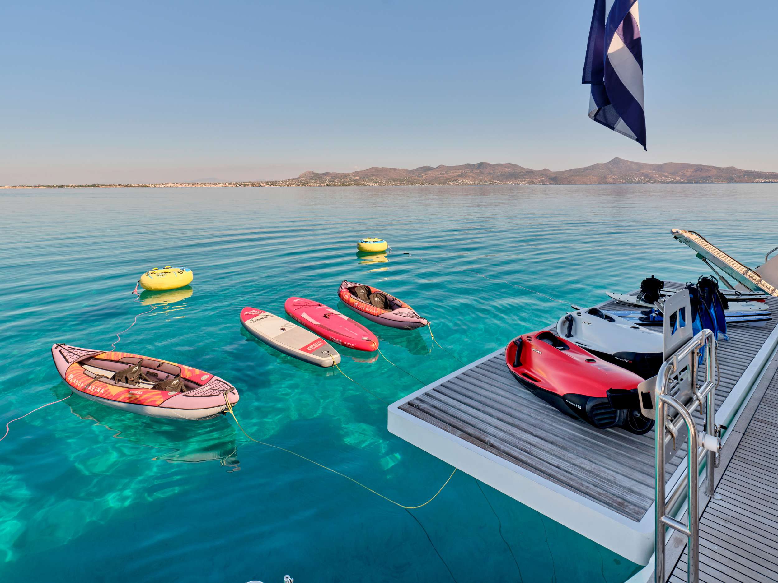 A deck overlooking crystal clear blue water with inflatable boats, paddleboards, and water toys floating nearby; a Greek flag waves on the right, with distant hills visible across the calm sea.