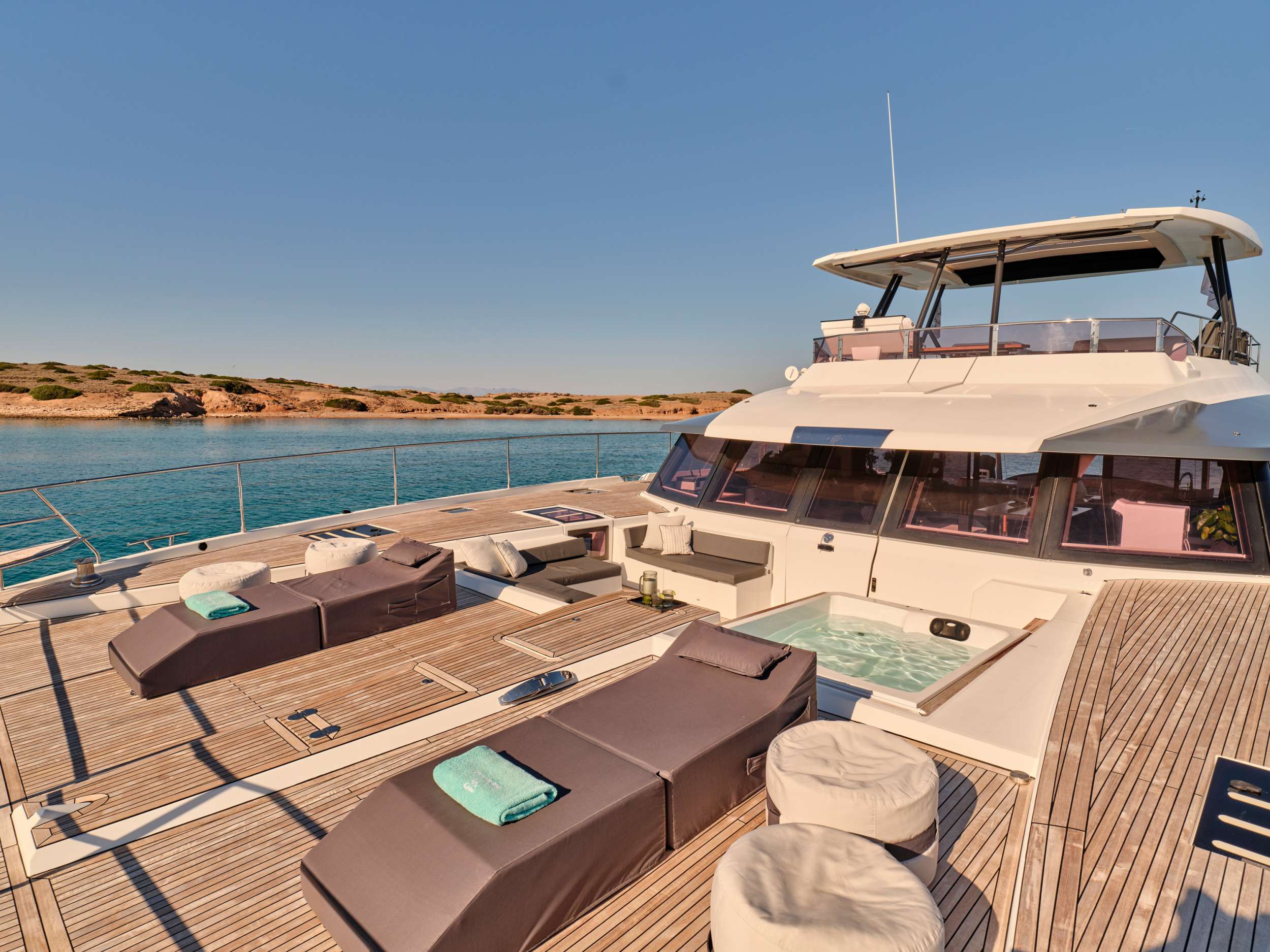 A luxury yacht deck with cushioned sun loungers, a hot tub, and round poufs overlooks calm turquoise water and a rocky shoreline under a clear blue sky.