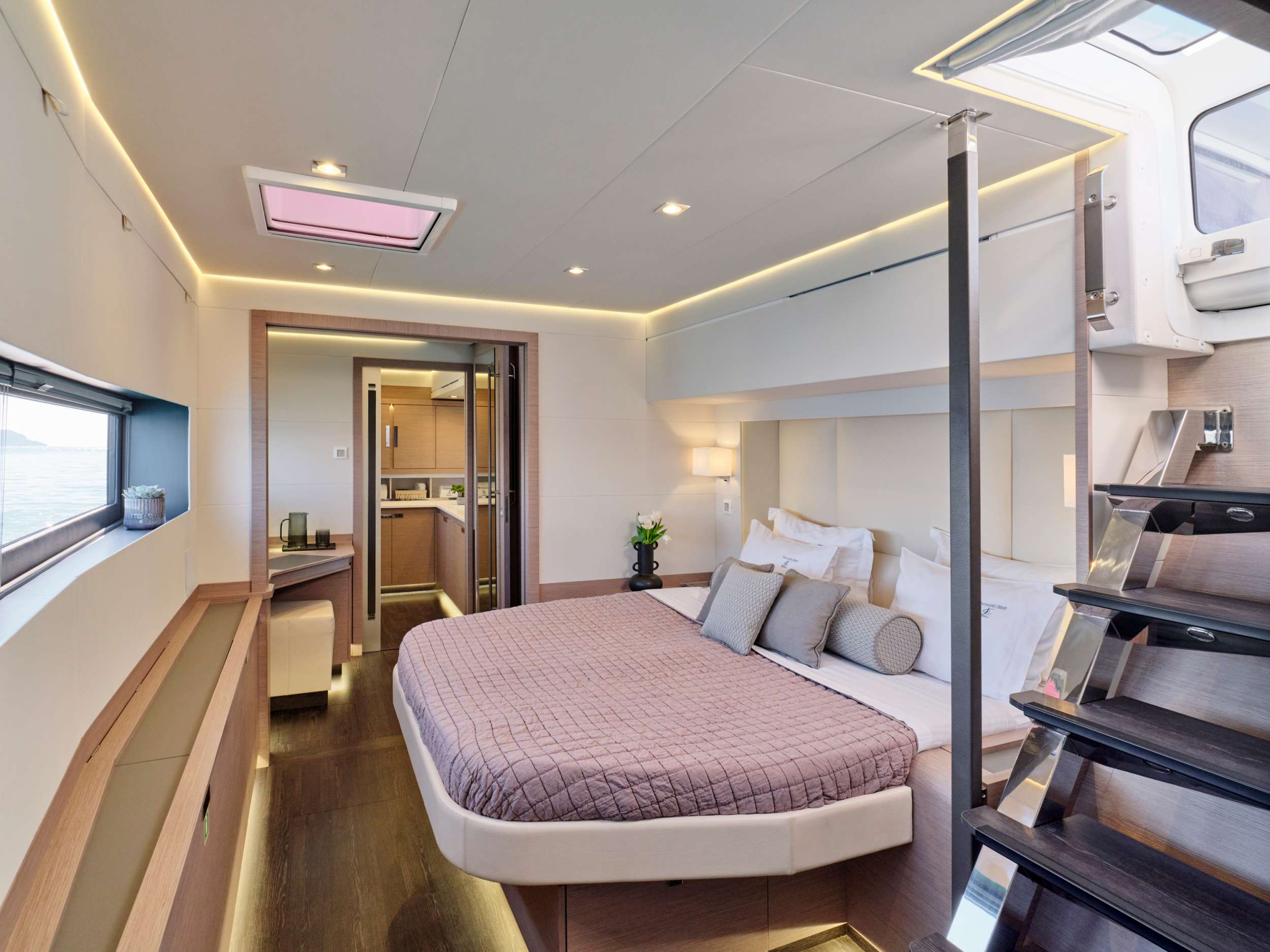 A modern yacht bedroom with a large bed, light wood accents, soft lighting, decorative pillows, and windows offering a view of the water. Stairs lead up to a hatch, and the room features sleek, minimalist décor.