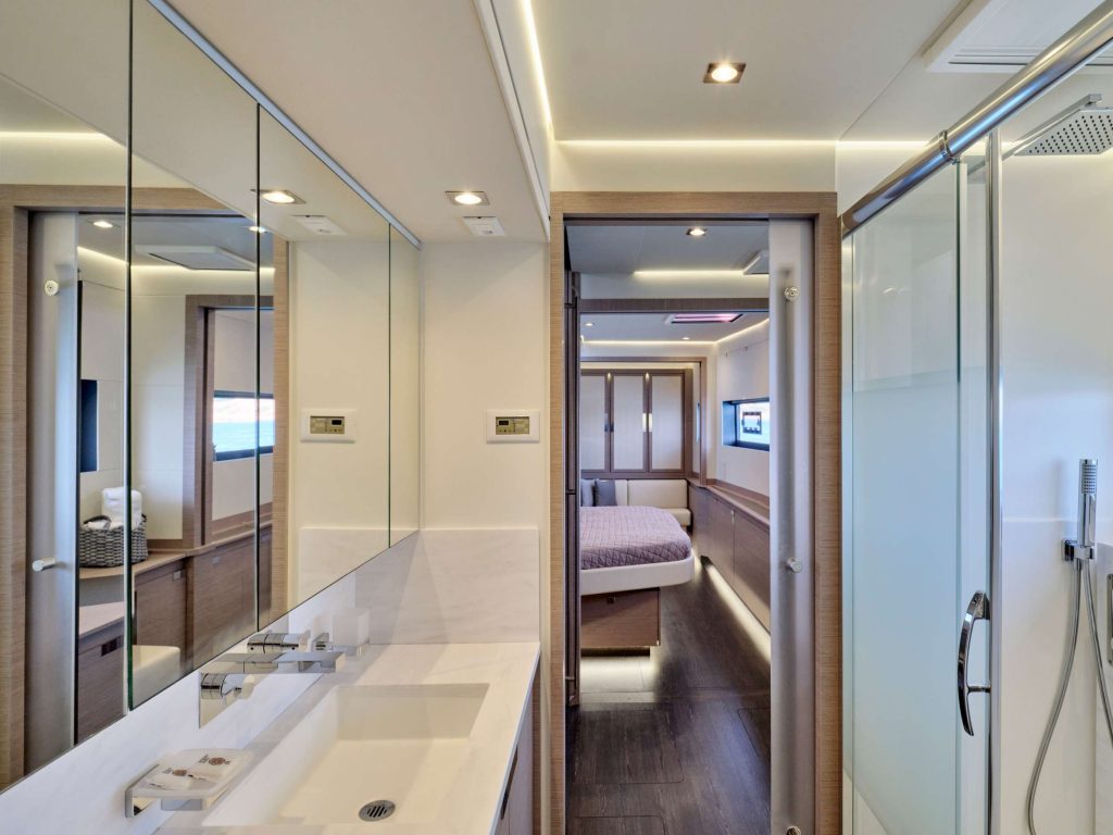 Modern bathroom with a double sink, large mirrors, glass shower, and light wood accents, leading to a bedroom with a bed, built-in storage, and TV in a sleek, minimalist interior design.
