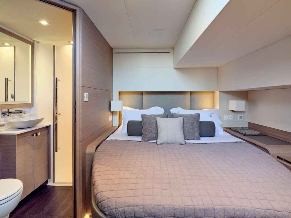 Modern bedroom on a yacht featuring a large neatly-made bed with neutral bedding and multiple pillows, built-in nightstands, soft lighting, and an adjacent compact bathroom with a sink and mirror.