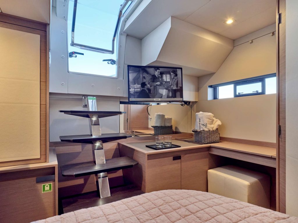 Modern yacht cabin with light wood decor, a bed with a pink quilt, a wall-mounted TV showing a black-and-white program, floating stairs leading to a skylight, and a compact desk area with towels and a small cushioned stool.
