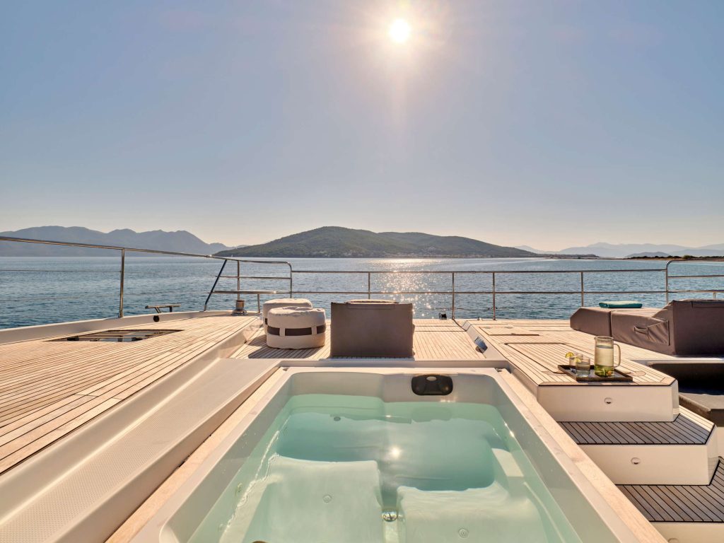 A luxury yacht deck with a hot tub overlooking calm blue waters and distant hills under a bright sun, with lounge chairs and a drink on a side table.