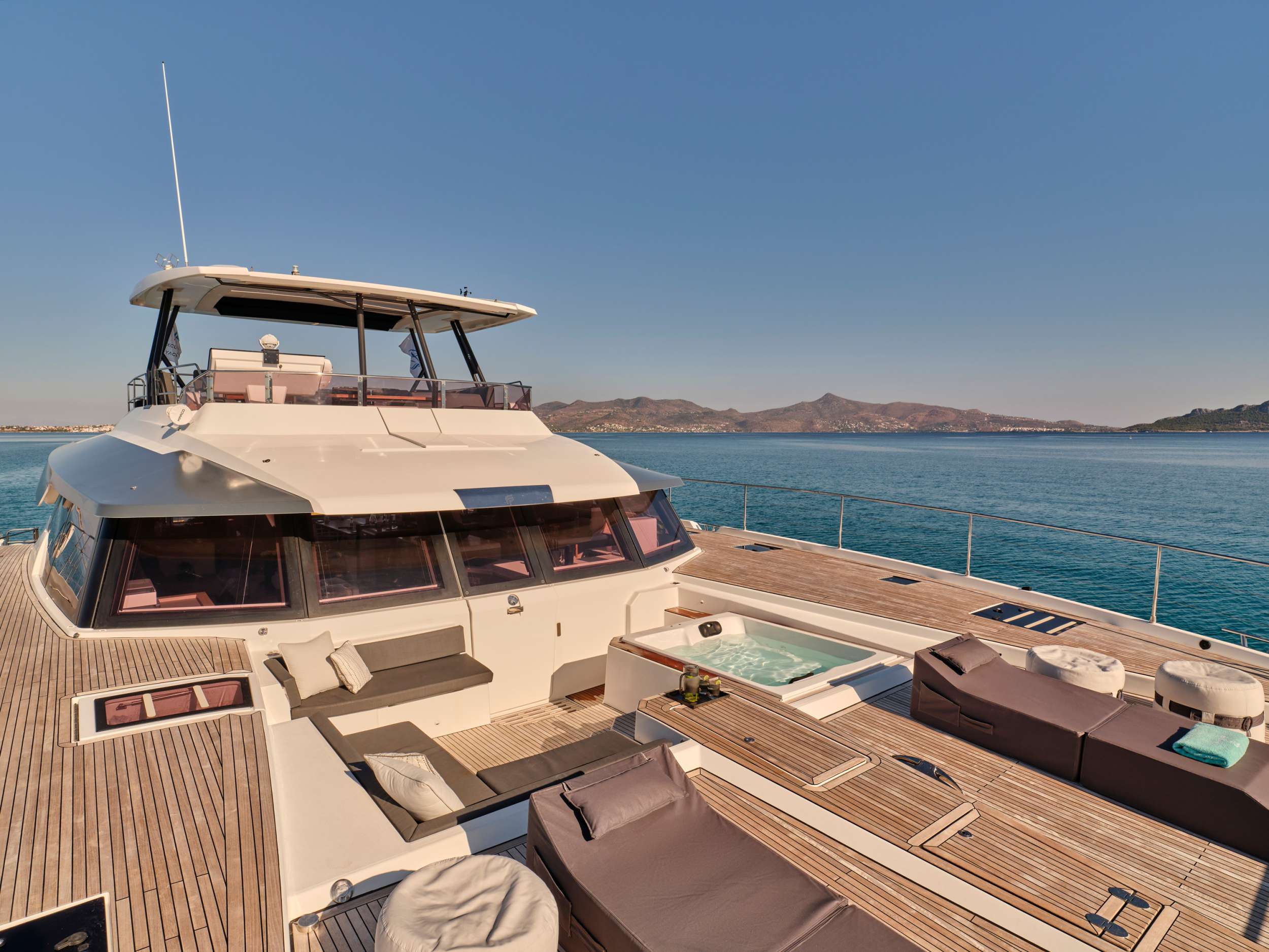 A luxurious yacht deck with ample seating, a jacuzzi, and sun loungers, floating on calm blue water with mountains visible in the distance under a clear sky.