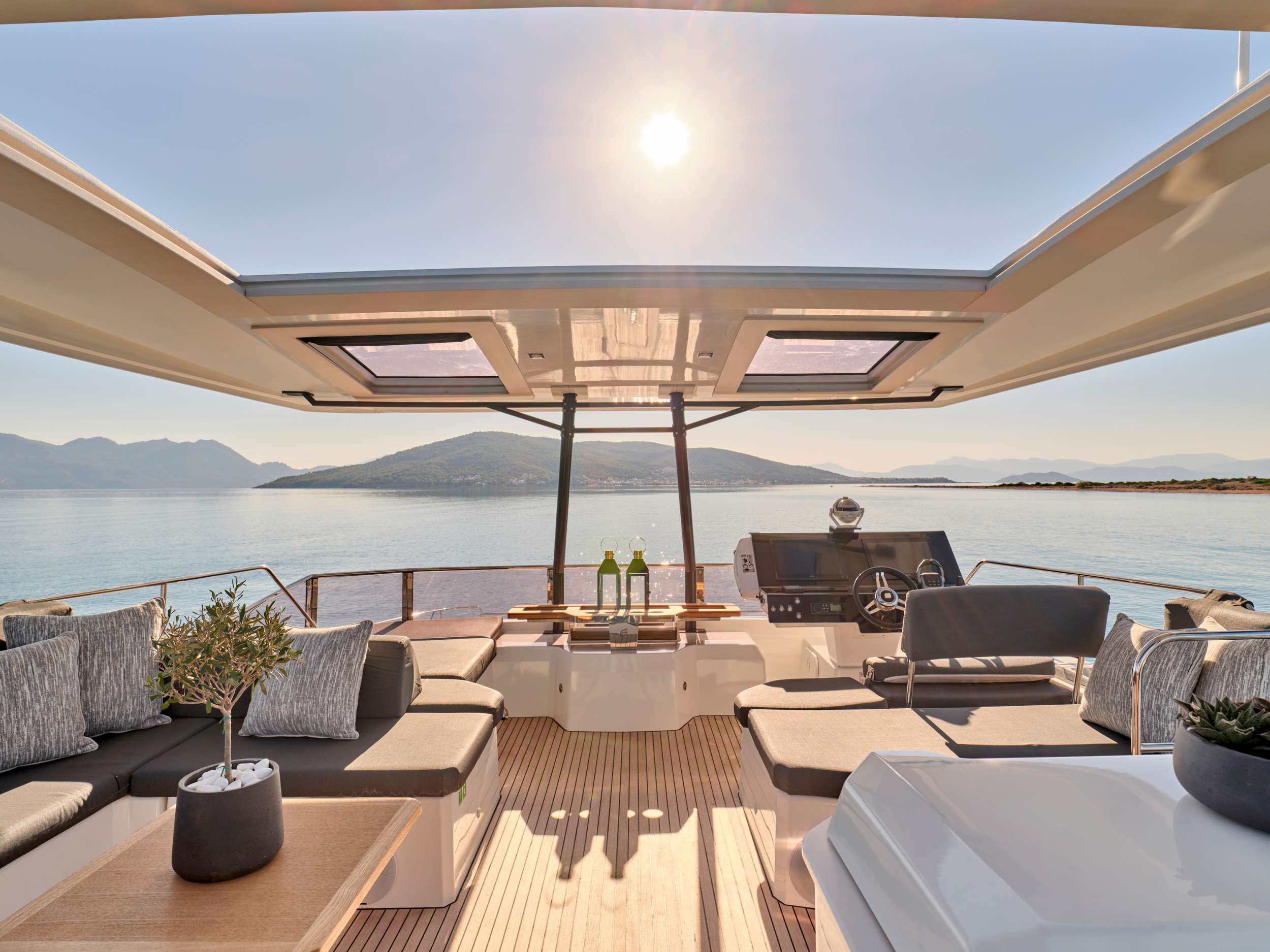 A luxurious yacht deck with cushioned seating, a small table set with wine and glasses, and scenic views of calm water and distant mountains under a bright sun.
