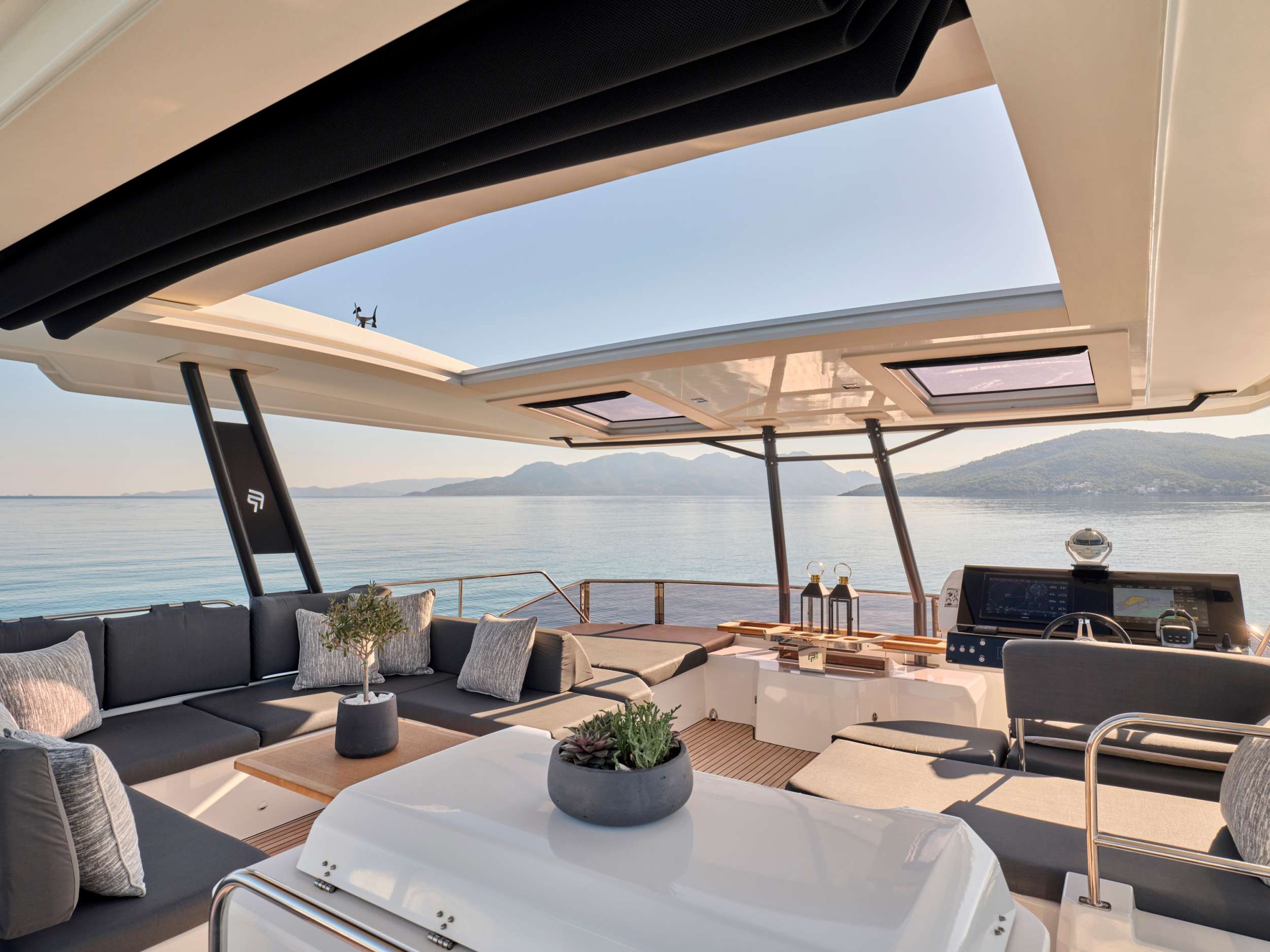 A luxurious yacht deck with modern seating, tables, and potted plants, shaded by a retractable roof. The yacht is on calm water with mountains visible in the background under a clear sky.
