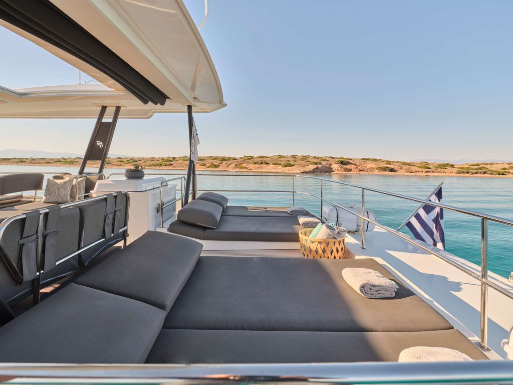 Sun loungers with gray cushions and rolled towels on a yacht deck, overlooking calm blue water and a distant coastline, with a Greek flag displayed at the stern under a clear sky.