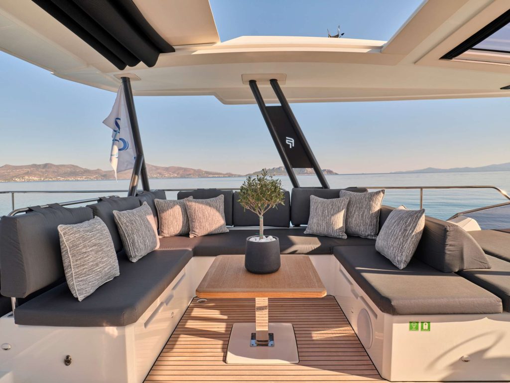 Modern yacht deck with a large L-shaped sofa featuring grey and white cushions, a small potted plant on a wooden table, and calm sea with distant mountains in the background under a clear sky.