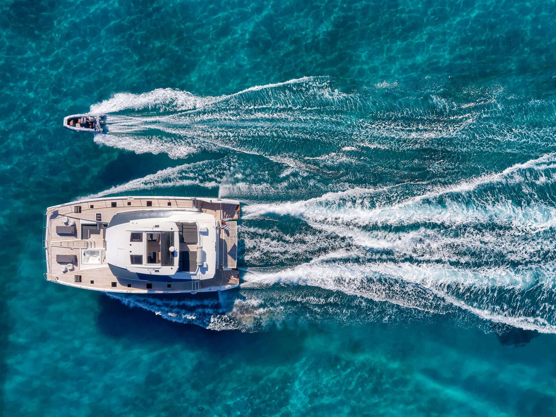 Aerial view of a large white yacht sailing in clear blue water, leaving trails of white foam, with a smaller speedboat following closely behind.