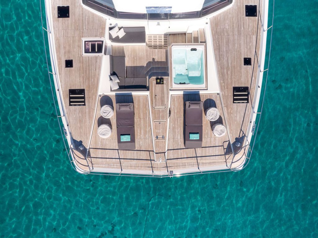 Aerial view of a yacht deck with lounge chairs, a hot tub, and seating area, floating on clear turquoise water.
