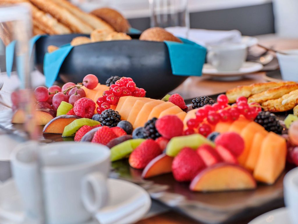 A close-up of a breakfast table with a platter of assorted fresh fruits, including melon, berries, grapes, kiwi, and apple slices, with bread and coffee cups in the background.
