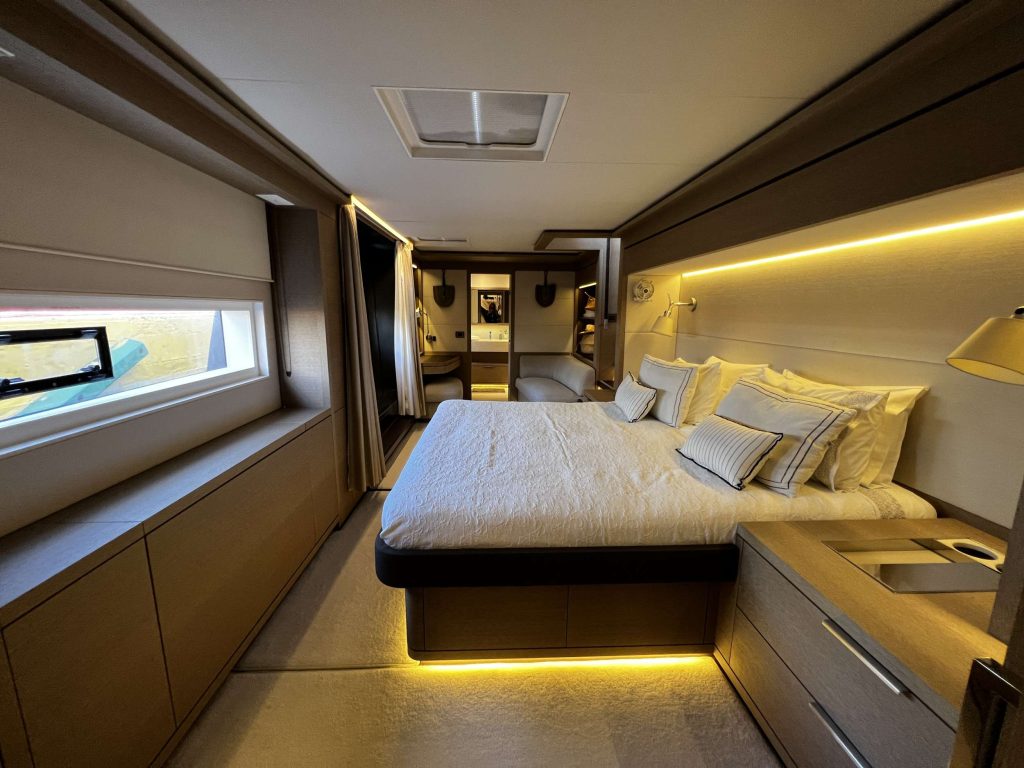 A modern, well-lit bedroom with a large bed, plush white bedding, wood-paneled walls, built-in nightstands, and soft under-bed lighting. A window runs along one side and an open doorway leads to a bathroom.