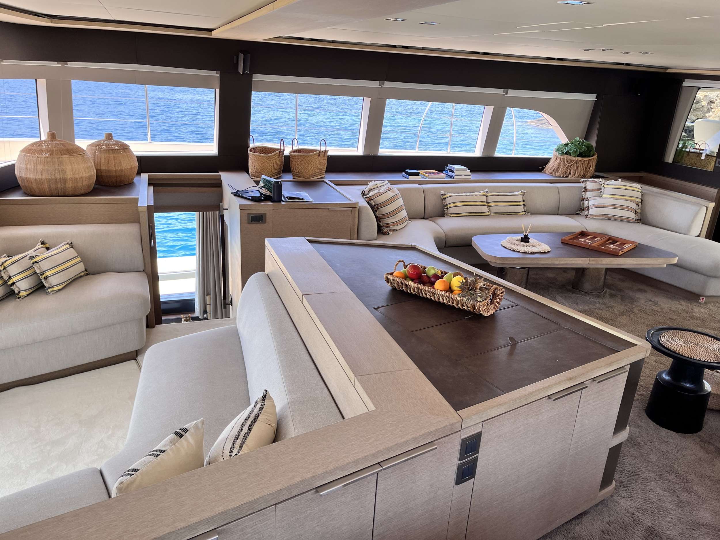 Modern yacht interior with large windows, beige sectional sofas, striped pillows, wicker baskets, and a table with a fruit basket. The ocean is visible outside the windows, creating a bright and relaxing atmosphere.