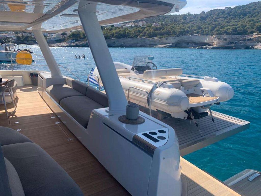 A luxury yacht deck with gray seating, a small table with a cup, and a white inflatable boat on a platform at the stern, floating on clear blue water near a rocky shoreline.