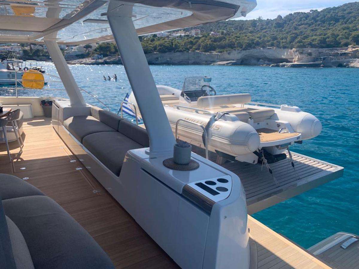 A luxury yacht deck with gray seating, a small table with a cup, and a white inflatable boat on a platform at the stern, floating on clear blue water near a rocky shoreline.