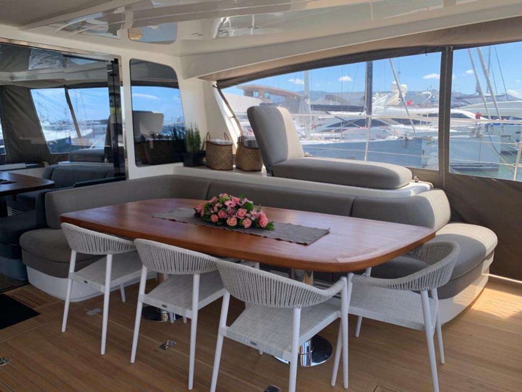 A yacht interior with a wooden dining table, six white chairs, and a gray cushioned bench. A pink flower arrangement is on the table. Large windows show a marina and other boats outside.