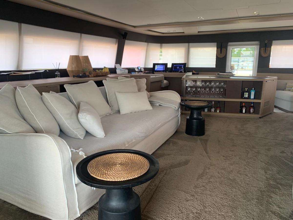 A spacious yacht interior with a large beige sofa, several white pillows, two small round tables, a bar area with drinks and glassware, and large windows letting in natural light.