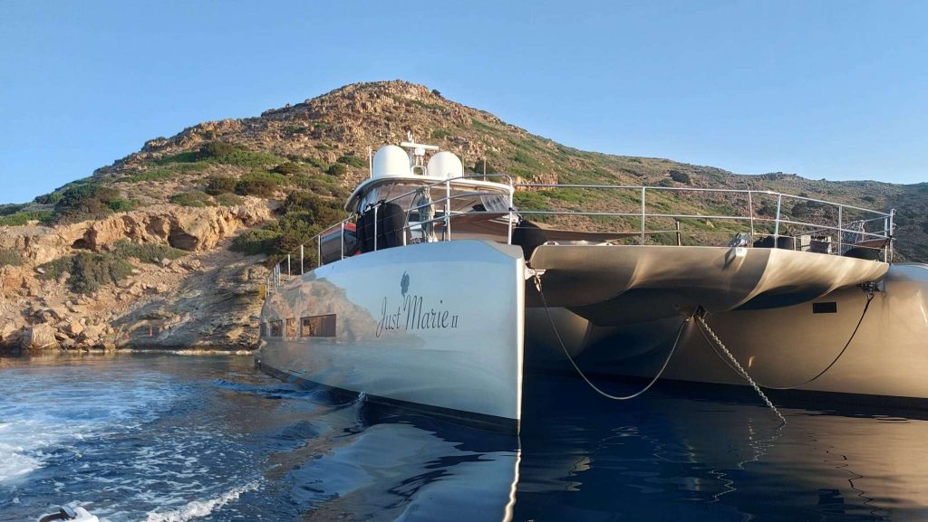 A modern catamaran yacht named Just Marie 2 is anchored near a rocky shoreline with a hill in the background, under a clear blue sky. The yacht reflects in the calm water.