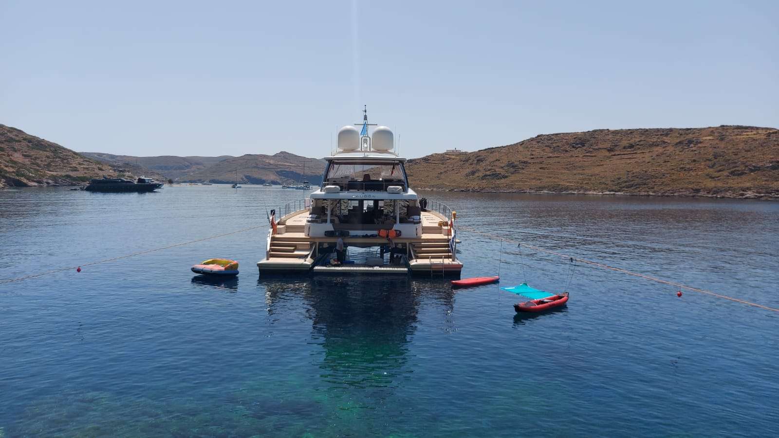 A large yacht is anchored in calm, clear blue water near a rocky shoreline. Small inflatable rafts float nearby, and distant hills are visible under a clear sky.