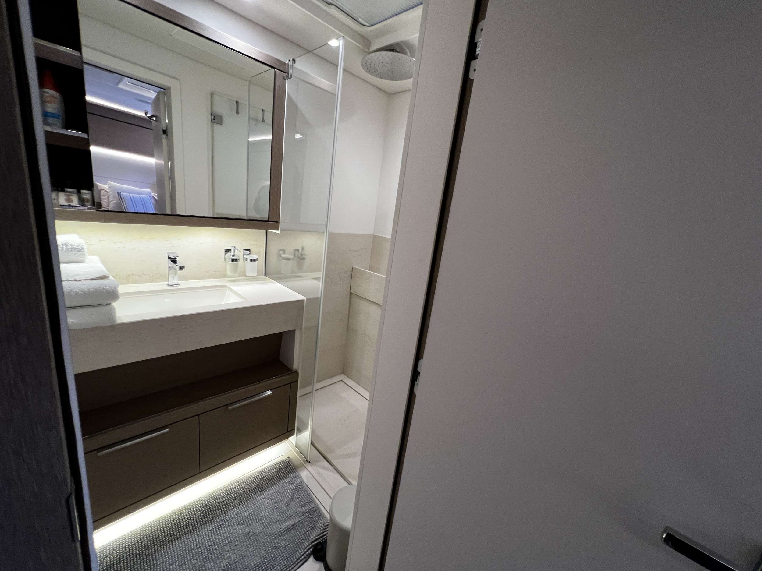 Modern bathroom with a large mirror above a sink, towels neatly stacked on the counter, a glass-enclosed shower, shelves with toiletries, and soft lighting under the vanity. The door is partially open.