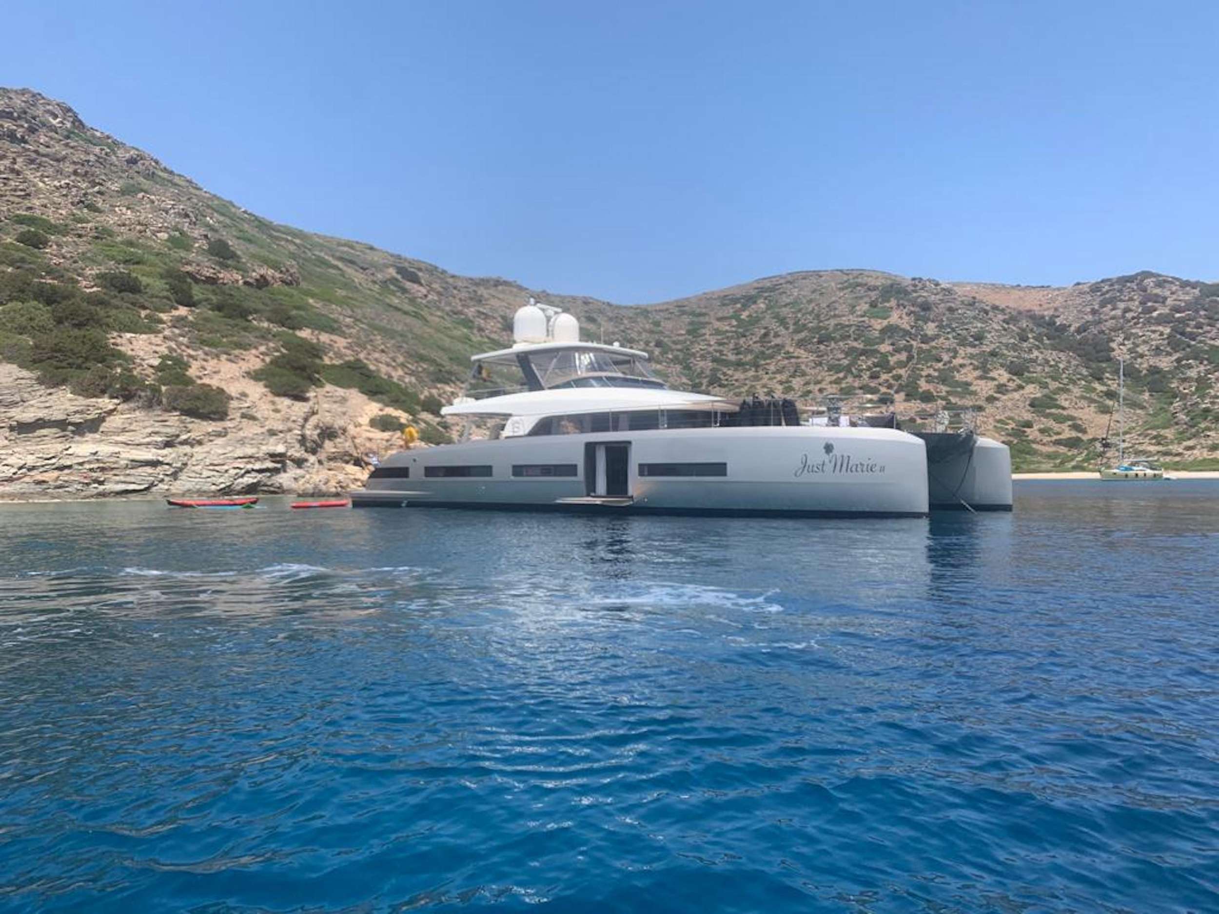 A large white yacht named Just Marie is anchored near a rocky, green hillside and clear blue water under a bright sky. Several small boats float nearby.