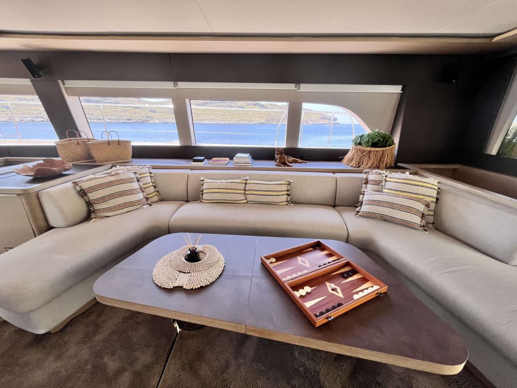 A modern yacht interior with a light gray L-shaped sofa, striped pillows, a table with a decorative tray and a backgammon set, baskets, and large windows overlooking the water and rocky shoreline.