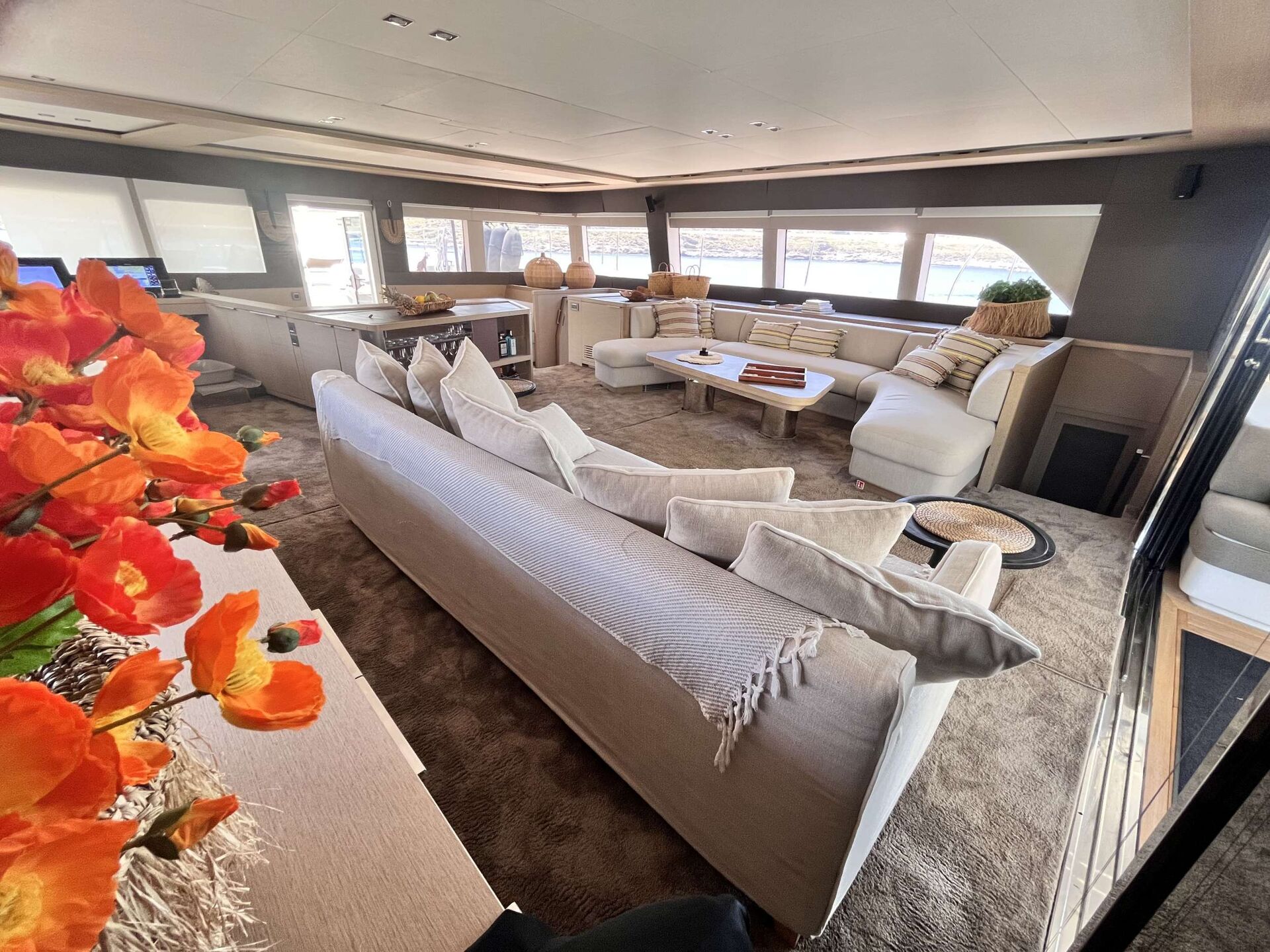 A spacious yacht lounge with beige sofas, light cushions, a white table, and large windows. Orange flowers in the foreground add a vibrant touch to the cozy, modern interior.