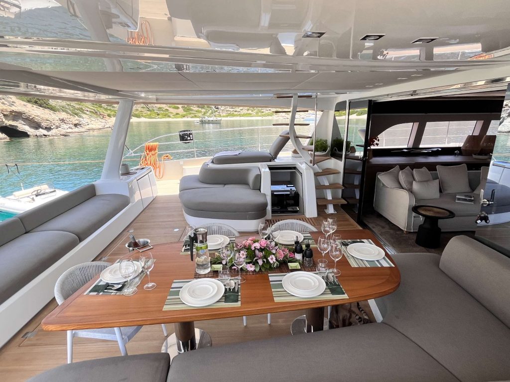 A luxury yacht’s deck set for a meal, with a wooden table, dishes, wine, and flowers; gray cushioned seating surrounds the area. The yacht overlooks calm water and rocky green shores in the background.