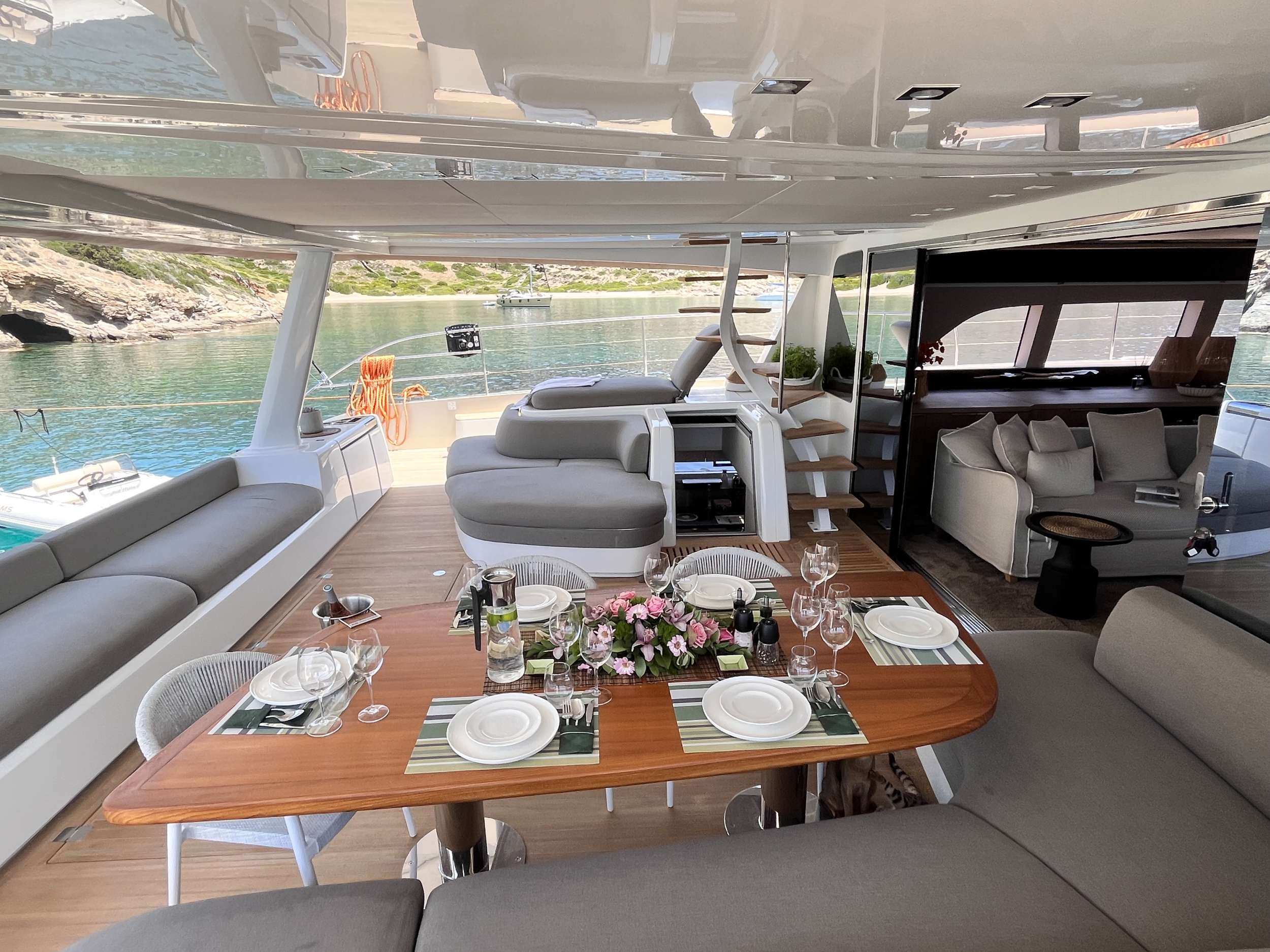 A luxury yacht’s deck set for a meal, with a wooden table, dishes, wine, and flowers; gray cushioned seating surrounds the area. The yacht overlooks calm water and rocky green shores in the background.