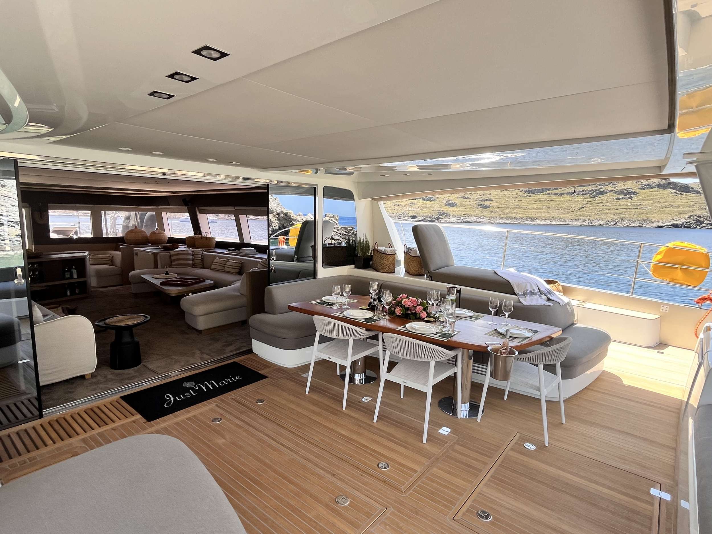 Luxurious yacht interior with a dining table set for six on a wood deck, cushioned seating, and an open view of calm water and rocky shore in the background. The indoor lounge is stylishly furnished.